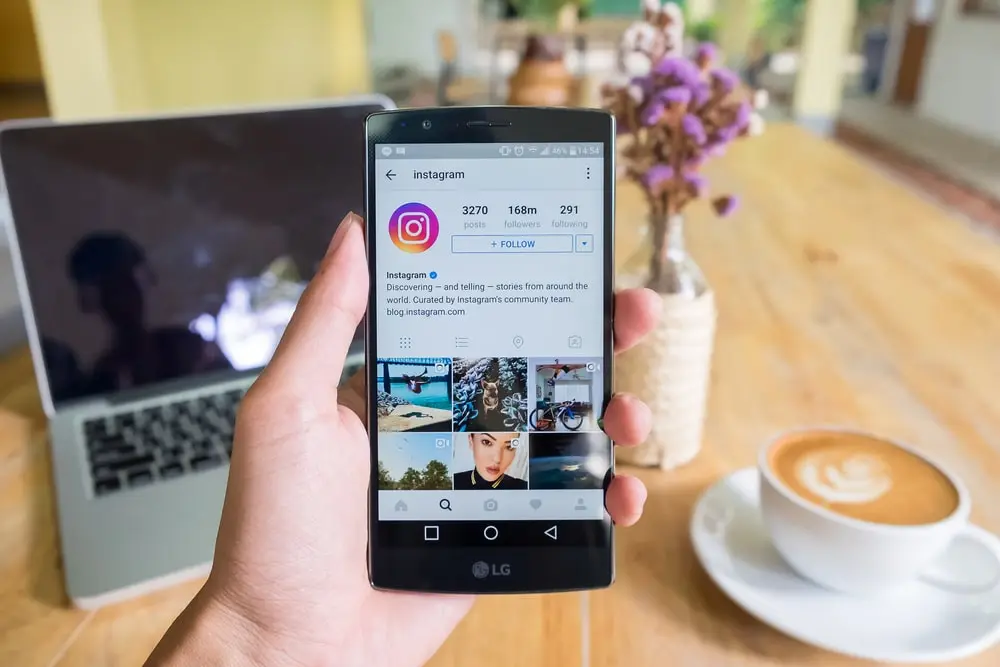 Person holding a smartphone displaying Instagram’s official profile, with a laptop, coffee cup, and vase of flowers on a wooden table in the background.